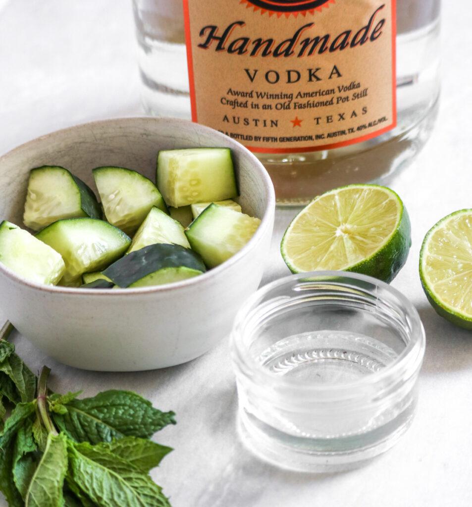 ingredients for a cucumber mint martini on a white marble counter: vodka, cucumber, mint, lime, and simple syrup.